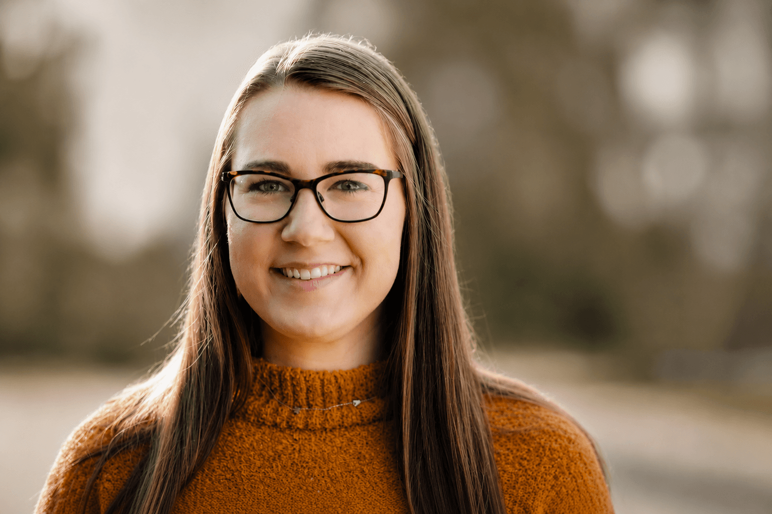 Smiling woman with long brown hair and tortoiseshell glasses wearing an orange sweater outdoors.